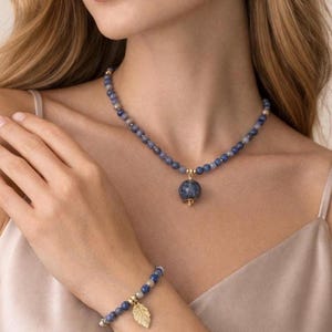 Puede incluir: Un conjunto de joyas con un collar de cuentas, una pulsera y pendientes. El collar tiene un colgante grande y redondo. La pulsera tiene un dije de hoja dorada. Las cuentas son azules y doradas. Los pendientes son pequeñas cuentas azules redondas.