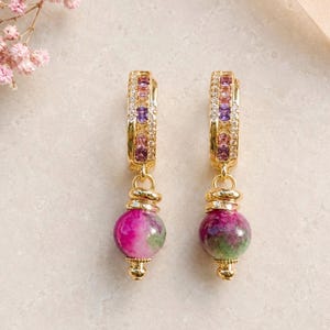 Puede incluir: Un par de pendientes de aro dorados con una hilera de pequeñas piedras preciosas cuadradas de color púrpura y rosa, flanqueadas por cristales transparentes. Cada pendiente tiene una cuenta esférica colgante con un patrón jaspeado en magenta, púrpura y verde.