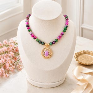 Puede incluir: Un collar de cuentas con piedras redondas alternas en color magenta y verde oscuro. El collar incluye un colgante de tono dorado con un borde de filigrana intrincado que rodea una piedra central ovalada de color morado claro y detalles claros.