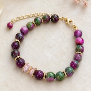 Può includere: Un braccialetto di perline con pietre rotonde e levigate in tonalità viola intenso e verde bosco, accentuato da piccoli distanziatori in metallo dorato e un ciondolo cilindrico incastonato con cristalli rosa chiaro taglio baguette e chiusura a moschettone.