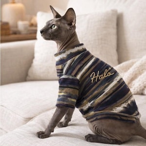 Peut inclure: Un chat Sphynx gris portant un pull rayé avec le mot "Halo" brodé en or. Le pull présente des rayures horizontales dans les tons de bleu, marron et blanc. Le chat est assis sur un canapé blanc.