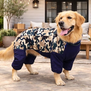 Op de afbeelding: Een Golden Retriever draagt een camouflage en marineblauwe hondenjas. De jas heeft een patroon van groene, bruine en beige vormen. De hond staat op een stenen terras.