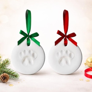 May include: Two white, round Christmas ornaments with a paw print design. Each ornament has a ribbon for hanging, one green and one red. The ornaments are festive and suitable for pet lovers.