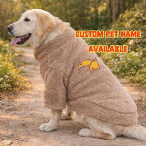 Peut inclure: Un golden retriever porte un pull-over marron et duveteux pour chien avec un motif jaune et orange. Le texte "CUSTOM PET NAME AVAILABLE" est imprimé sur le pull. Le chien est assis sur un chemin de terre.