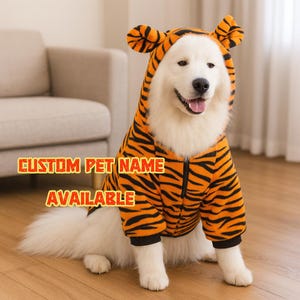 May include: A white dog wearing an orange and black tiger-striped pet costume with a hood and ears. The costume has a zip closure. Text on the image reads "CUSTOM PET NAME AVAILABLE".