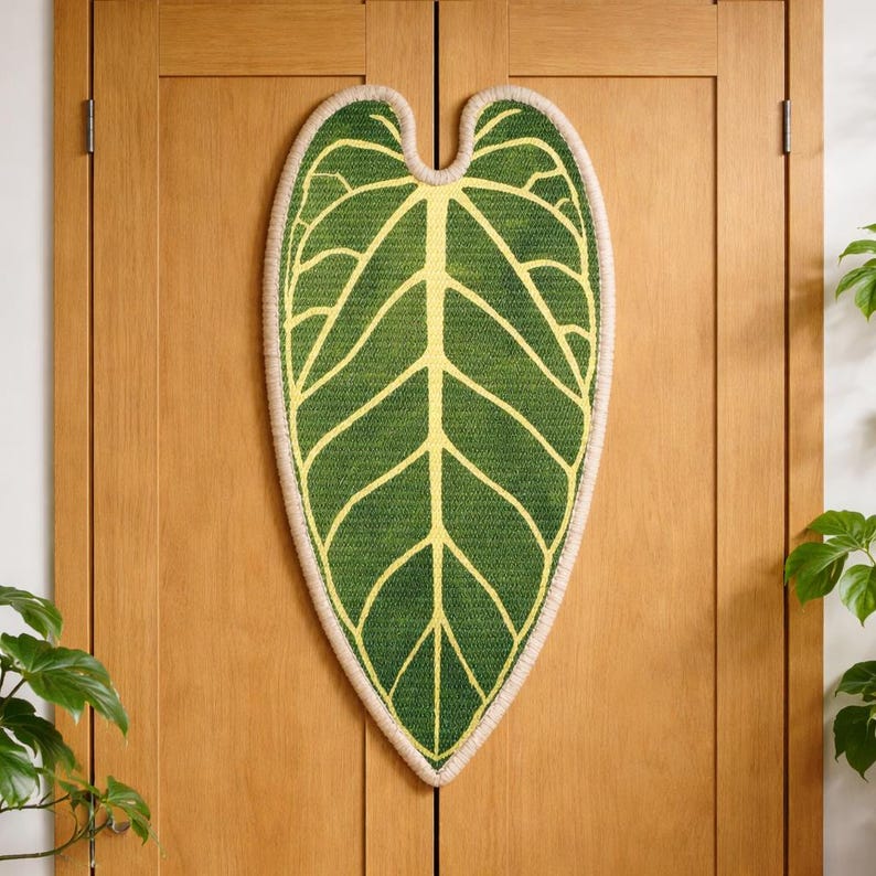 May include: A green leaf-shaped doormat with yellow veins and a beige border. The mat is made of woven material and is hanging on a wooden door. The design resembles an Anthurium leaf.