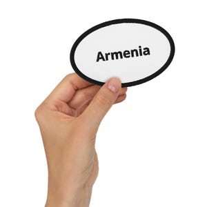 May include: An oval patch with a white background and a black border. The word "Armenia" is printed in black, centered on the patch. The patch is held by a hand against a white background.