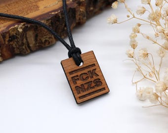 May include: A rectangular wooden pendant hanging from a black cord. The pendant features the engraved text FCK NZS arranged in two lines, framed by horizontal lines at the top and bottom. The wood has a natural brown grain and a smooth finish.