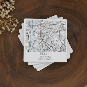 Può includere: Una pila di sottobicchieri quadrati bianchi con una mappa nera e azzurra di Praga, Repubblica Ceca. Il nome della città e le coordinate sono stampati sotto. I sottobicchieri sono su una superficie di legno scuro.