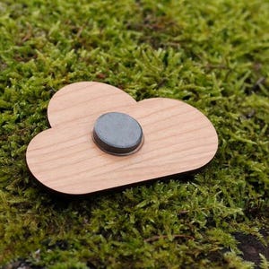 May include: A wooden cloud-shaped magnet with a dark gray circular magnet in the center. The magnet is light brown and sits on a bed of green moss.