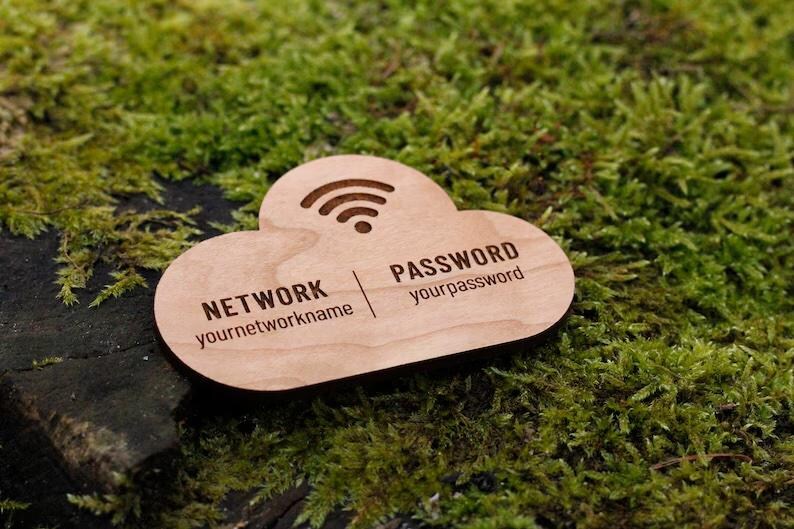 May include: A wooden cloud-shaped sign with engraved text for network and password information. The sign includes a Wi-Fi symbol and is set against a backdrop of green moss.