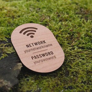 May include: A wooden WiFi password sign with a rounded shape. The sign features a WiFi symbol at the top and the words "NETWORK" and "PASSWORD" with placeholders for network name and password. The sign is set against a backdrop of green moss.