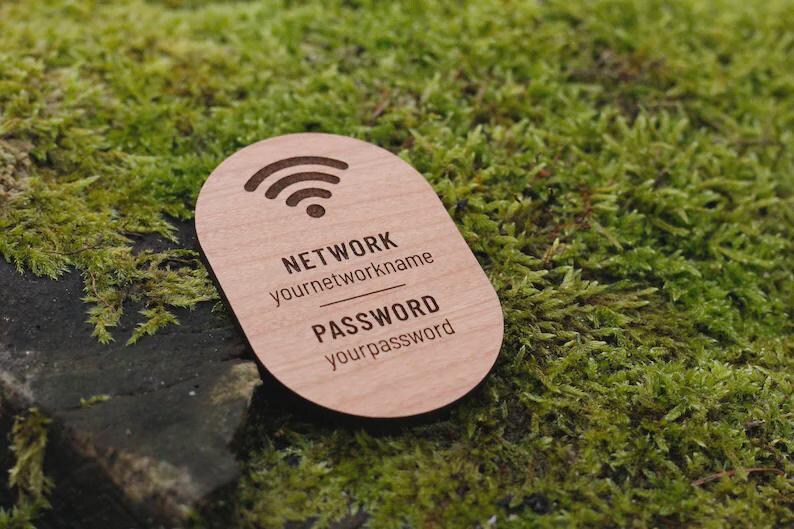 May include: A wooden WiFi password sign with a rounded shape. The sign features a WiFi symbol at the top and the words "NETWORK" and "PASSWORD" with placeholders for network name and password. The sign is set against a backdrop of green moss.