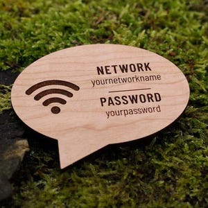 May include: A wooden speech bubble-shaped sign with engraved text and a Wi-Fi symbol. The sign reads "NETWORK yournetworkname" and "PASSWORD yourpassword". The sign is light brown and sits on a bed of green moss.