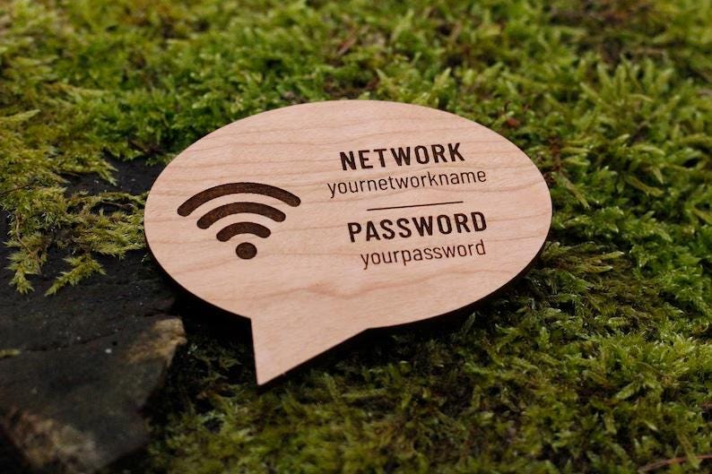 May include: A wooden speech bubble-shaped sign with engraved text and a Wi-Fi symbol. The sign reads "NETWORK yournetworkname" and "PASSWORD yourpassword". The sign is light brown and sits on a bed of green moss.