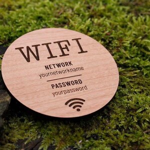 May include: A round wooden WiFi sign with engraved text. The sign includes the word "WIFI", network name, password, and a WiFi symbol. The sign is light brown and rests on a bed of green moss.