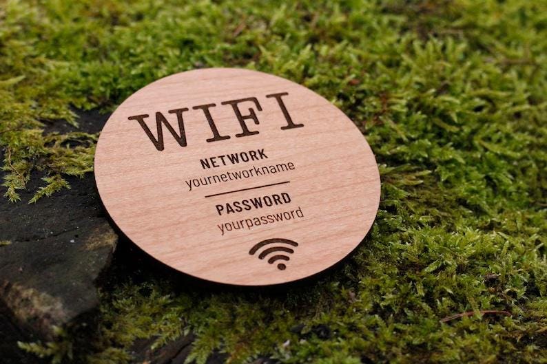 May include: A round wooden WiFi sign with engraved text. The sign includes the word "WIFI", network name, password, and a WiFi symbol. The sign is light brown and rests on a bed of green moss.