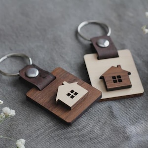 May include: Two wooden keyrings with house-shaped cutouts. One keyring is a dark brown square with a light brown house, and the other is a light brown rectangle with a dark brown house. Each has a silver key ring and a brown leather strap.