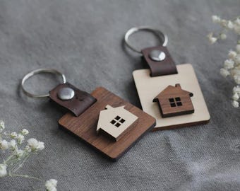 May include: Two wooden keychains with house-shaped cutouts. One keychain is a dark brown square with a light brown house, and the other is a light brown rectangle with a dark brown house. Each has a silver key ring and a brown leather strap.