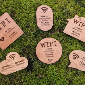 May include: Wooden WiFi password signs in various shapes, including a square, oval, house, cloud, circle, and speech bubble. Each sign has the word "WIFI", network name, and password printed on it. The signs are on a bed of green moss.