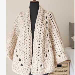 Peut inclure: Cardigan en crochet crème, sans fermeture, présenté sur un mannequin. Le tricot épais présente un tissage lâche avec des détails ajourés. Les manches sont aux trois quarts, et le style général est décontracté et confortable. Le cardigan est de couleur neutre.