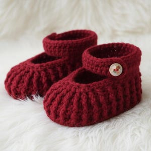 May include: A pair of handmade, burgundy crochet baby shoes. The shoes have a Mary Jane style with a strap and button closure. The shoes are made of a thick yarn and sit on a white, fluffy surface.