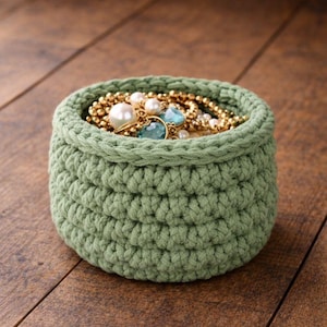 May include: A sage green, crocheted basket filled with jewelry. The basket is round with a textured stitch pattern. The jewelry includes gold chains, pearl necklaces, and rings with blue and clear gemstones. The basket sits on a wooden surface.