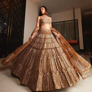 May include: A gold-toned lehenga, choli, and dupatta set, heavily embellished with intricate detailing. The skirt has a geometric design. This formal outfit is suitable for special occasions, such as weddings or other celebrations.