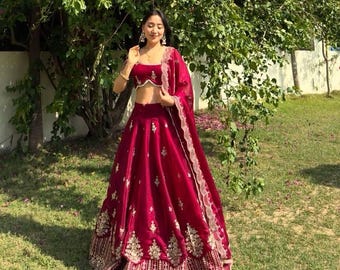 May include: A deep red, two-piece formal outfit with gold embroidery. The outfit includes a crop top, a long skirt, and a matching shawl. The ensemble is worn outdoors in front of a tree with pink flowers.