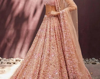 May include: A light pink, embellished lehenga with a matching choli and dupatta. The skirt is full and adorned with pearls and floral embroidery. The choli has a sweetheart neckline. The dupatta is sheer and drapes over the shoulder.