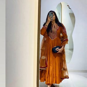 Peut inclure: Une robe orange à manches longues avec des broderies complexes autour de l'encolure et des manches. La robe a une jupe fluide et est associée à un châle transparent orné. Une petite pochette bleu foncé est tenue.