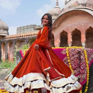 Peut inclure: Une robe orange vif à manches longues et une jupe ample, ornée d'argent. La robe est portée devant un bâtiment orné avec un textile à motifs roses et dorés.