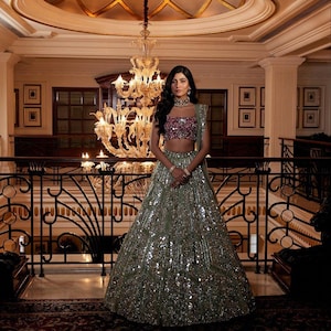 May include: A woman in a formal gown stands in a grand hall. The gown is a light green colour and covered in sequins. The top is a pink and purple sequined crop top. A large chandelier hangs above.