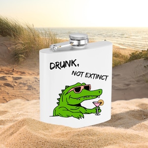May include: A white metal flask with a silver screw-top lid. The front features a graphic of a green alligator wearing pink sunglasses and holding a pink cocktail with an orange slice. The text reads: DRUNK, NOT EXTINCT in black letters.