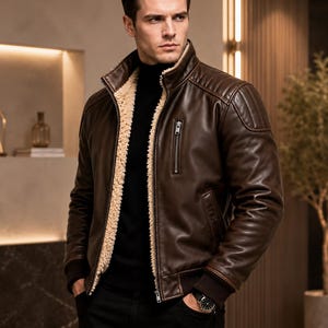 May include: A dark brown leather jacket featuring a cream-colored sherpa lining, a high collar, a vertical zippered chest pocket, and ribbed cuffs. The jacket is worn over a black turtleneck sweater and paired with black trousers.