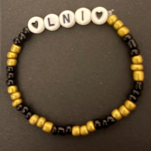 May include: A beaded bracelet spelling "LNIO" with white letter beads. The bracelet has a pattern of gold and black beads. This handmade bracelet is a stylish accessory, suitable for everyday wear.