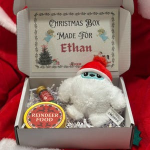 May include: A Christmas gift box containing a plush yeti toy in a red hat, a small bottle, and a round container labelled "Reindeer Food". The box is personalised with the text "Christmas Box Made For Ethan".