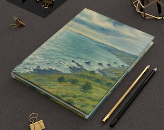 Puede incluir: Un diario de tapa dura con una pintura de acuarela de un paisaje costero. La portada presenta una vista del océano, los acantilados y el cielo en tonos de azul, verde y amarillo. Lápices dorados y negros y clips dorados están junto al diario.