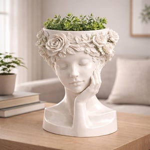 May include: A white ceramic planter shaped like a woman's head, adorned with a floral crown of roses. The planter holds green plants. The woman's face has closed eyes and a hand resting on her cheek. The planter sits on a wooden table.