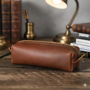 Puede incluir: Un estuche de lápices de cuero marrón con cierre de cremallera. El estuche rectangular tiene una parte superior redondeada y un pequeño lazo en un extremo. El estuche está sobre una superficie de madera, con libros y una lámpara de escritorio en el fondo.