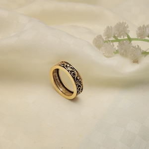 May include: A gold-toned ring with an intricate black filigree design. The ring is set against a soft, off-white background, with a sprig of white flowers in the upper right corner. The ring's band features a repeating pattern of swirling lines.