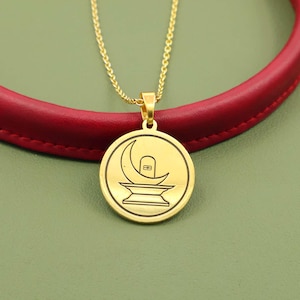 May include: Gold-colored pendant necklace featuring a circular pendant with a black etched design. The design includes a crescent moon, a stylized shape, and a base. The necklace is on a gold chain, set against a red and green background.