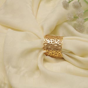 May include: A gold-colored napkin ring with an openwork design of interconnected organic shapes. The ring is wrapped around a cream-colored fabric, creating a decorative table setting. White flowers are in the background.