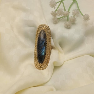 Op de afbeelding: Een goudkleurige ring met een ovale, iriserende blauwgrijze steen. De ring heeft een bewerkte, sierlijke rand en ligt op een crèmekleurige stof. Een takje witte bloemen staat op de achtergrond.
