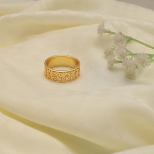May include: A gold-colored ring with a repeating pattern of bear figures around the band. The ring is set against a cream-colored fabric, with a sprig of white flowers in the background. The ring appears to be a size 7.