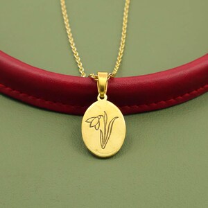 May include: Gold-toned necklace featuring an oval pendant with a floral design. The pendant hangs from a delicate chain. The background includes a red leather band and a green surface.