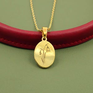 May include: A gold-colored pendant necklace with an oval charm featuring a delicate floral design. The pendant hangs from a thin, gold chain. The background includes a red leather-like material and a green surface.