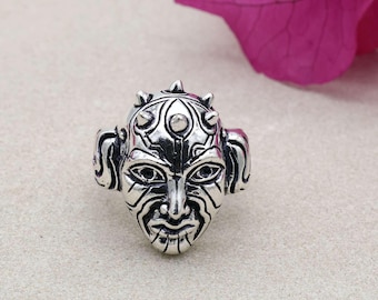 May include: A silver ring with a detailed face design. The ring features an intricate face with pointed features and a textured surface. The design suggests a unique, artistic style. The ring is likely a statement piece, suitable for adding a distinctive touch to an outfit.
