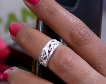 May include: A silver ring with an openwork design featuring intricate floral and scroll patterns. The ring is worn on a finger with red nail polish. The ring's design is visible, showcasing its delicate craftsmanship and style.