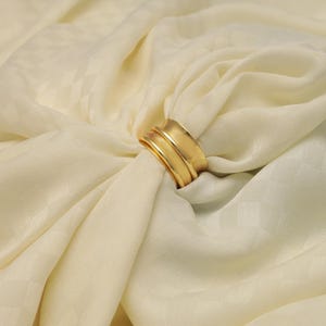 Peut inclure: Une bague dorée avec un design à plusieurs couches, posée sur un tissu ivoire. La surface de la bague reflète la lumière, soulignant sa texture lisse. Le tissu présente un motif de losanges subtil.